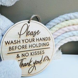 May include: A round wooden sign with the text "Please wash your hands before holding & no kisses Thank you!" in a cursive font. The sign is hanging from a string and is in front of a grey stuffed animal and a rainbow-coloured yarn wreath.