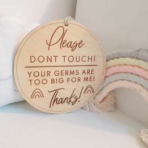 May include: A round wooden sign with the text "Please Don't Touch! Your germs are too big for me! Thanks!" in a brown font. The sign is decorated with a rainbow design and is hanging from a string.