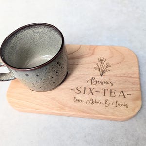 May include: A grey speckled ceramic mug with a brown rim and handle rests on a wooden serving board. The board is engraved with the words "Bessa's - SIX-TEA - love. Abbie & Louis" and a floral design. A tea set.