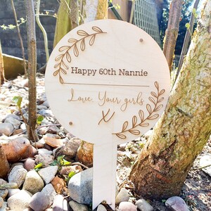 May include: A wooden sign with a leafy design around the edges. The sign says "Happy 60th Nannie Love Your Girls X".