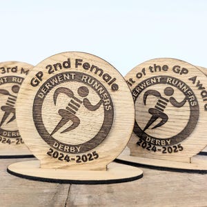 May include: Wooden awards with a circular design, featuring the text "GP 2nd Female" and "Derwent Runners Derby 2024-2025." The awards have a running figure graphic and are made of light-coloured wood.