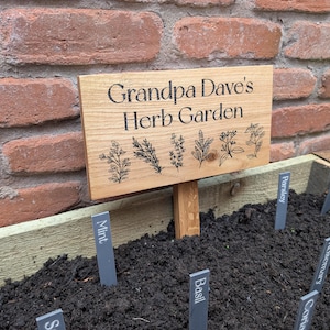 May include: A wooden sign reading "Grandpa Dave's Herb Garden" with herb illustrations. The sign is in a garden bed with markers for herbs like thyme, mint, and basil. A brick wall provides the backdrop for this garden scene.