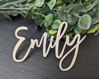 May include: A wooden sign with the name "Emily" written in a cursive font. The sign is light brown and has a natural wood finish.