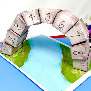 May include: A papercraft bridge made of nine numbered cubes, each with a different number from 1 to 9. The bridge is arched over a blue river on a green and brown landscape.