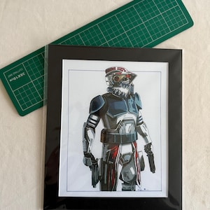 Peut inclure: Illustration encadrée d'un personnage en casque et armure, tenant deux armes. Le personnage est principalement gris, blanc et rouge. Le cadre est noir et un tapis de découpe vert est en arrière-plan.