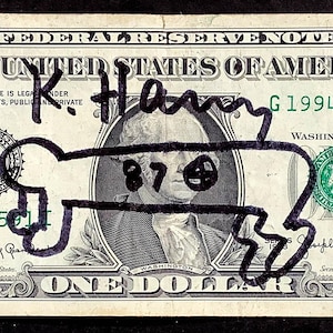 K&ouml;nnte beinhalten: Ein Ein-Dollar-Schein mit einer schwarzen Markierung, die eine Knochenform &uuml;ber dem Portr&auml;t von George Washington darstellt. Auf der Rechnung steht mit schwarzem Marker "K. Hany". Die Seriennummer der Rechnung lautet G19943591I.