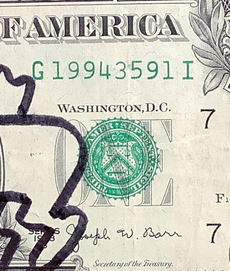 K&ouml;nnte beinhalten: Nahaufnahme eines Ein-Dollar-Scheins mit dem Wort "AMERICA" oben. Die Seriennummer "G19943591 I" ist sichtbar. Der Schein hat ein gr&uuml;nes Siegel und den Text "WASHINGTON, D.C."