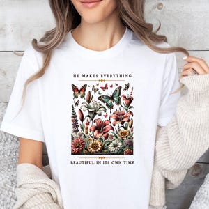 Op de afbeelding: Wit T-shirt met een bloemen- en vlinderontwerp. De tekst "He makes everything beautiful in its own time" staat op het T-shirt gedrukt.