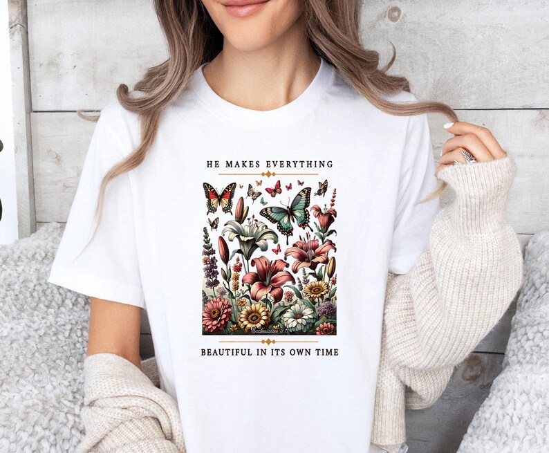 Op de afbeelding: Wit T-shirt met een bloemen- en vlinderontwerp. De tekst "He makes everything beautiful in its own time" staat op het T-shirt gedrukt.