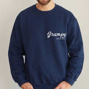 May include: A navy blue crewneck sweatshirt featuring the white text Grumpy SINCE 2023 printed on the upper left chest area. The garment has long sleeves, a ribbed neckline, and a solid dark blue colour throughout the fabric design.