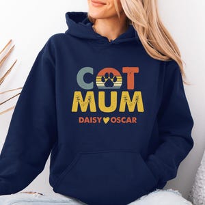 May include: A navy blue hooded sweatshirt featuring a graphic print with the text CAT MUM, a paw print icon, and the names DAISY and OSCAR separated by a heart symbol. The design uses a retro-style color palette of muted orange, yellow, and blue.