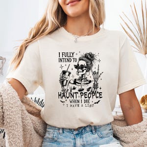 May include: Cream-colored t-shirt featuring a black graphic of a skeleton with sunglasses, holding a drink. The text reads "I FULLY INTEND TO HAUNT PEOPLE WHEN I DIE I HAVE A LIST."