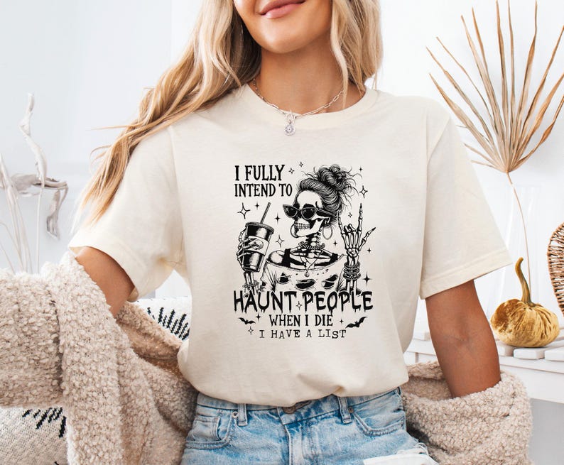 May include: Cream-colored t-shirt featuring a black graphic of a skeleton with sunglasses, holding a drink. The text reads "I FULLY INTEND TO HAUNT PEOPLE WHEN I DIE I HAVE A LIST."