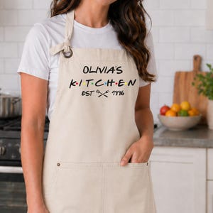 Puede incluir: Un delantal beige con el texto "OLIVIA'S KITCHEN EST 1996" en negro, con puntos de colores. El delantal tiene un bolsillo y una correa para el cuello. El delantal se usa en una cocina.