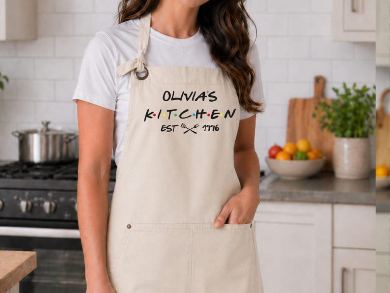 Puede incluir: Un delantal beige con el texto "OLIVIA'S KITCHEN EST 1996" en negro, con puntos de colores. El delantal tiene un bolsillo y una correa para el cuello. El delantal se usa en una cocina.