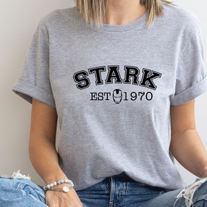 May include: A gray t-shirt with black text that reads "STARK EST 1970". A black and white image of Iron Man's mask is between the words "EST" and "1970".