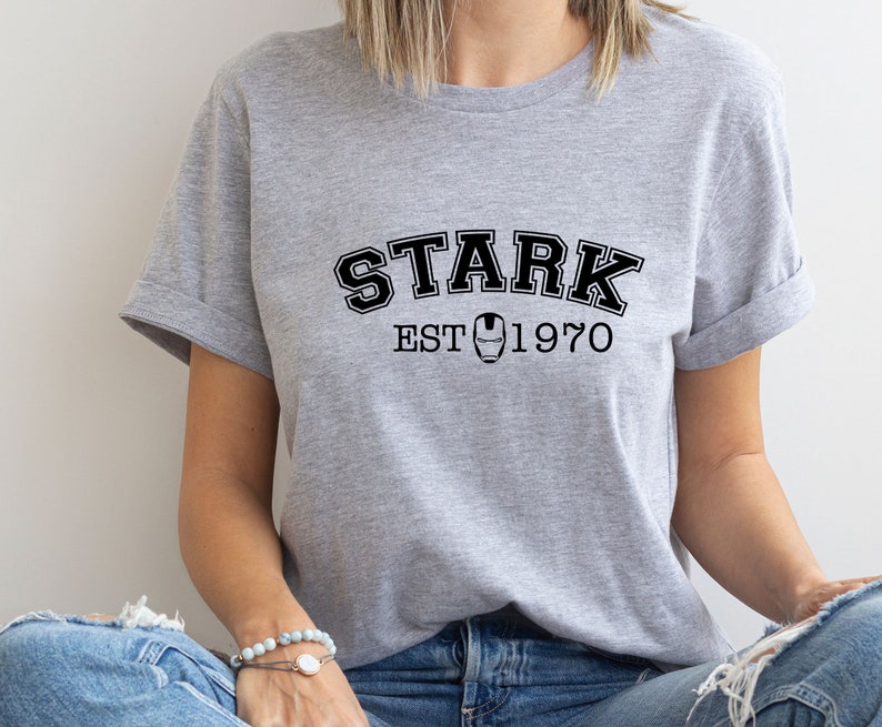 May include: A gray t-shirt with black text that reads "STARK EST 1970". A black and white image of Iron Man's mask is between the words "EST" and "1970".