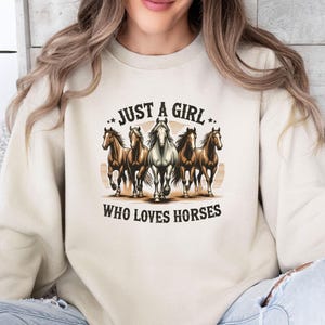 May include: A beige crewneck sweatshirt featuring a central graphic of five horses running forward. Above the horses, the text reads JUST A GIRL, and below the horses, the text reads WHO LOVES HORSES. The design uses brown, white, and black tones.