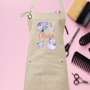 May include: A beige canvas apron with a pocket and a design of hairdressing tools and flowers. The text "Olivia" is printed on the apron.