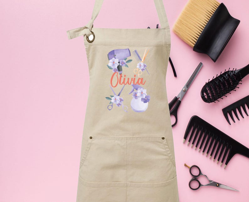 May include: A beige canvas apron with a pocket and a design of hairdressing tools and flowers. The text "Olivia" is printed on the apron.
