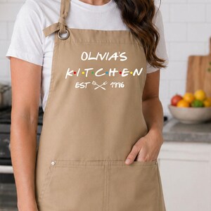 Puede incluir: Un delantal color beige con el texto blanco "Olivia's Kitchen", puntos de colores y el a&ntilde;o "1996". El delantal tiene un bolsillo y se lleva sobre una camiseta blanca. El entorno de la cocina incluye una estufa y una tabla de cortar.