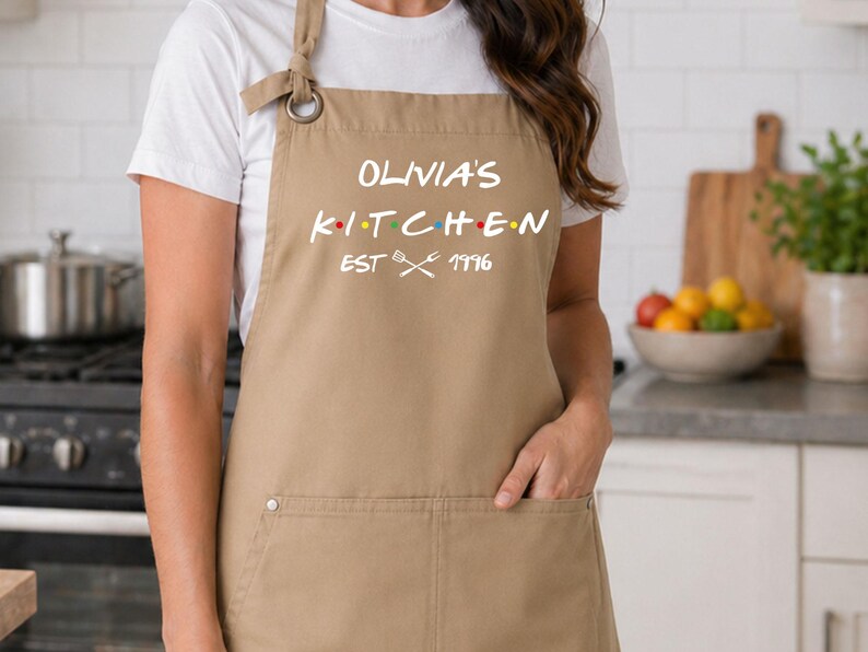 Puede incluir: Un delantal color beige con el texto blanco "Olivia's Kitchen", puntos de colores y el a&ntilde;o "1996". El delantal tiene un bolsillo y se lleva sobre una camiseta blanca. El entorno de la cocina incluye una estufa y una tabla de cortar.