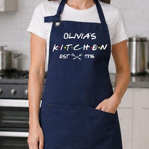 Puede incluir: Delantal azul marino con el texto blanco "OLIVIA'S KITCHEN", con puntos de colores entre las letras. El delantal tiene un bolsillo y el texto "EST 1996" con utensilios cruzados. Llevado sobre una camiseta blanca.