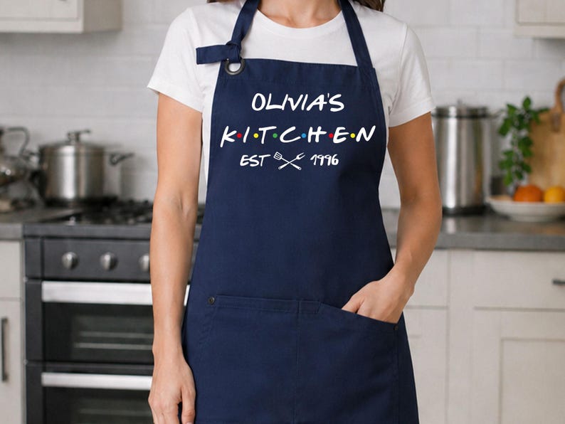 Puede incluir: Delantal azul marino con el texto blanco "OLIVIA'S KITCHEN", con puntos de colores entre las letras. El delantal tiene un bolsillo y el texto "EST 1996" con utensilios cruzados. Llevado sobre una camiseta blanca.