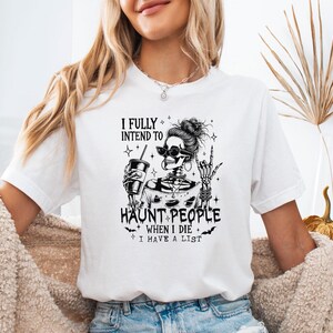 May include: White t-shirt with a black graphic of a skeleton wearing sunglasses and holding a drink. The text reads "I FULLY INTEND TO HAUNT PEOPLE WHEN I DIE I HAVE A LIST."