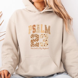 Peut inclure: Sweat à capuche beige avec une poche avant et un cordon de serrage. Le motif présente le mot "PSALM" et le chiffre "23" dans une police décorative, avec le texte "The Lord Is My Shepherd" en dessous.