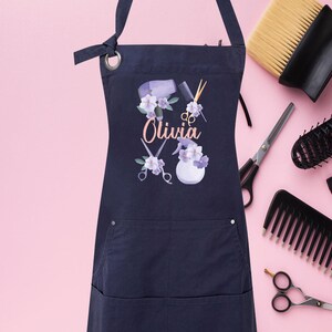 May include: A navy blue apron with a pocket and adjustable straps. The apron has a floral design with a hairdryer, scissors, and the name "Olivia" printed on it.