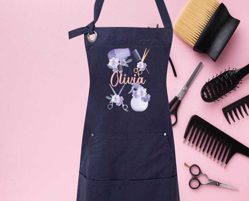 May include: A navy blue apron with a pocket and adjustable straps. The apron has a floral design with a hairdryer, scissors, and the name "Olivia" printed on it.