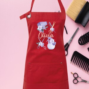 May include: Red apron with a pocket, featuring a design of hairdressing tools, flowers, and the name "Olivia".