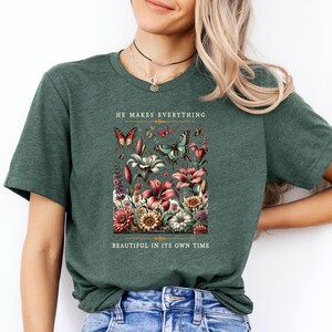 Op de afbeelding: Een groen T-shirt met een bloemenpatroon en vlinders. De tekst "He makes everything beautiful in its own time" staat op het shirt gedrukt.