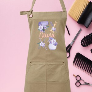 May include: A tan canvas apron with a pocket, featuring a floral design with hairdressing tools and the name "Olivia".