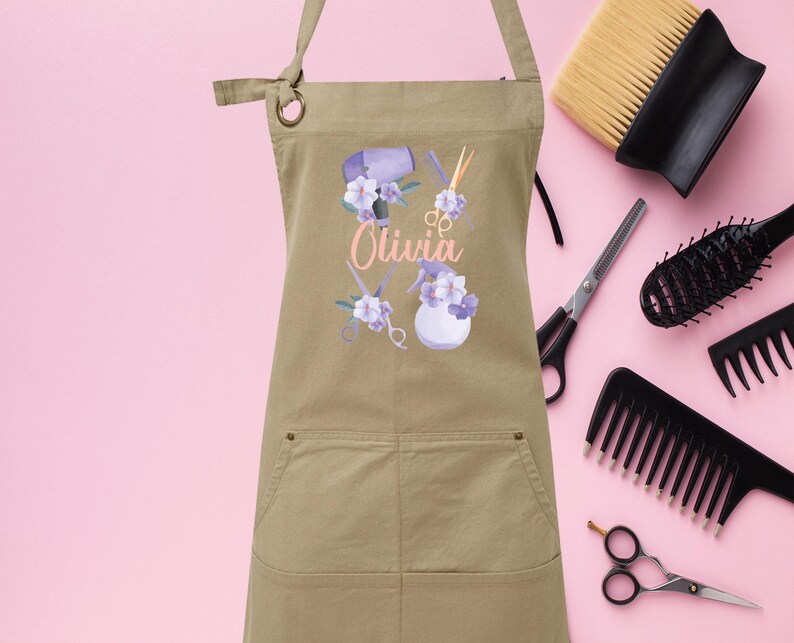 May include: A tan canvas apron with a pocket, featuring a floral design with hairdressing tools and the name "Olivia".