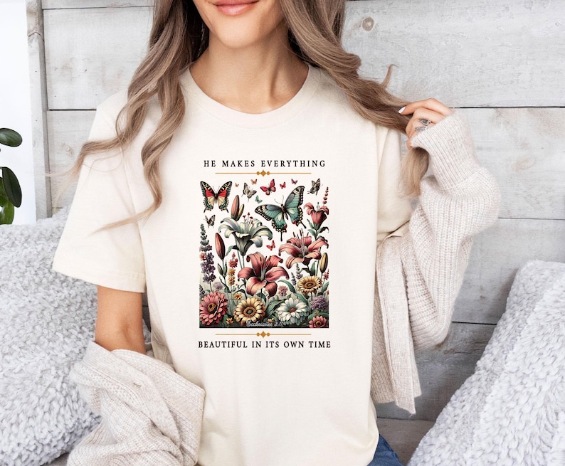 Op de afbeelding: Een cr&egrave;mekleurig T-shirt met een bloemenprint en vlinders. De tekst "He makes everything beautiful in its own time" staat op het shirt gedrukt.