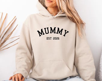 May include: A beige hooded sweatshirt featuring the black text MUMMY arched across the chest with the text EST 2026 printed directly below in a smaller font. The garment includes a front kangaroo pocket and long sleeves with ribbed cuffs.
