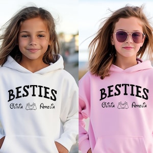 May include: Two girls wearing matching hoodies. One is white, the other pink. Both hoodies feature the word "BESTIES" in black, with the names "Olivia" and "Amelia" and a heart symbol printed below.
