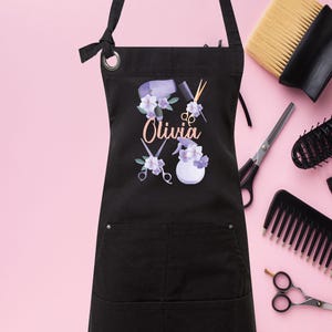 May include: Black apron with a pocket and adjustable straps. The apron has a design of hairdressing tools, flowers, and the name "Olivia".