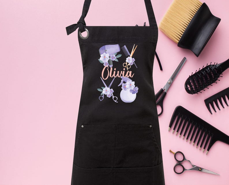 May include: Black apron with a pocket and adjustable straps. The apron has a design of hairdressing tools, flowers, and the name "Olivia".