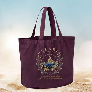 May include: A deep burgundy tote bag with two handles. The bag features a circular design with the text "VELARIS CITY OF STARLIGHT" and a mountain illustration. The quote "To the stars who listen and the dreams that are answered" is also printed on the bag.