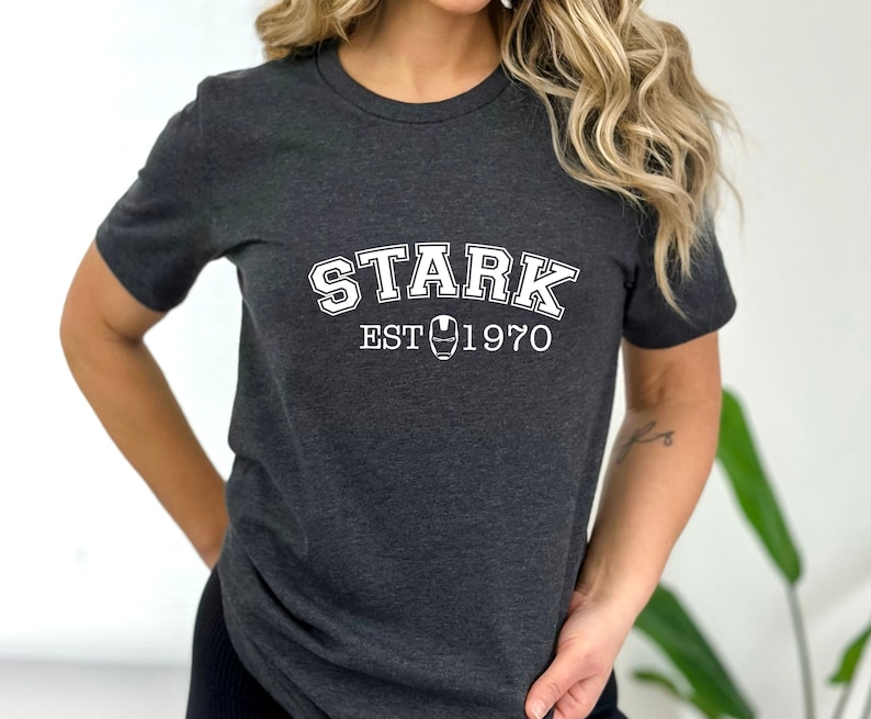 May include: A dark gray t-shirt with white text that reads "STARK EST 1970" with a stylized iron man mask in between the words "EST" and "1970".