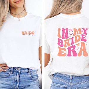 May include: White t-shirt with the text "BRIDE Amelia" in coral and pink on the front. The back of the shirt features the phrase "IN MY BRIDE ERA" in a retro font, with a ring graphic.