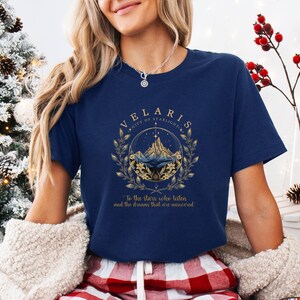 May include: A navy blue short-sleeve t-shirt featuring a gold and white graphic of mountains inside a circular frame with laurel branches. The text reads VELARIS CITY OF STARLIGHT and To the stars who listen and the dreams that are answered.