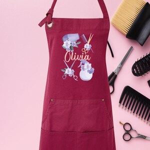 May include: A maroon apron with a pocket and adjustable straps. The apron has a design of hairdressing tools, flowers, and the name "Olivia".