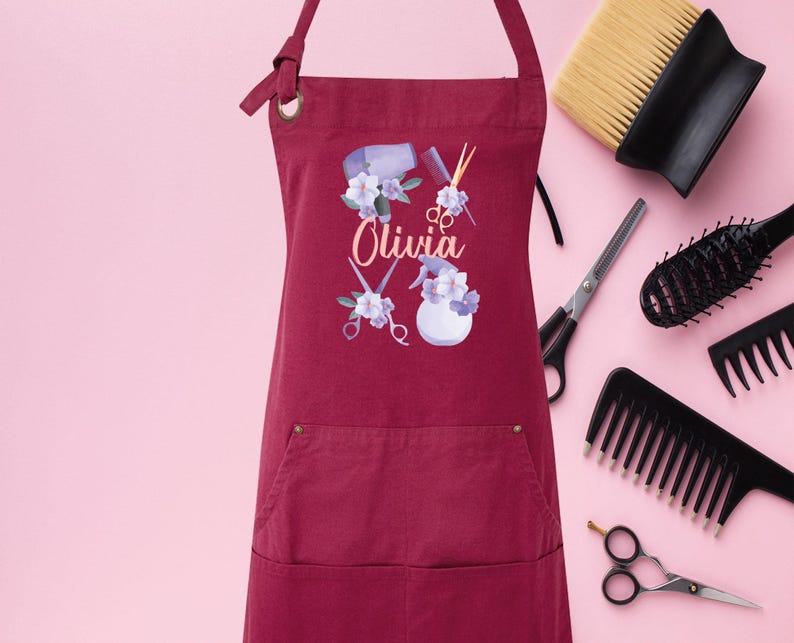 May include: A maroon apron with a pocket and adjustable straps. The apron has a design of hairdressing tools, flowers, and the name "Olivia".