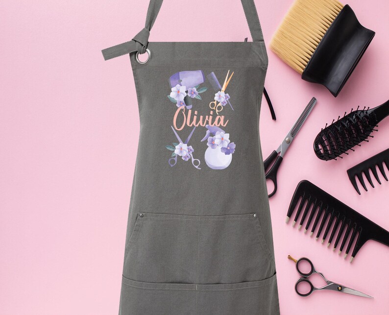 May include: A gray canvas apron with a pocket and adjustable straps. The apron has a floral design with hairdressing tools and the name "Olivia" printed on it.