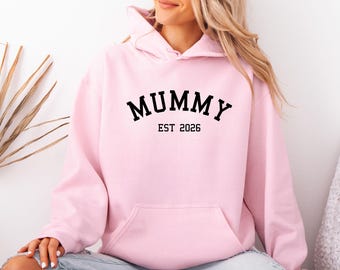 May include: A light pink hooded sweatshirt featuring a front kangaroo pocket and long sleeves. The text MUMMY EST 2026 is printed in a bold, black, arched serif font across the center of the chest, providing a clear and readable graphic design.