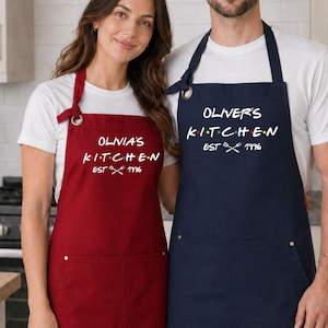 Puede incluir: Dos delantales, uno rojo y otro azul marino, con texto blanco. El delantal rojo dice "OLIVIA'S KITCHEN EST 1716". El delantal azul marino dice "OLIVER'S KITCHEN EST 1716". Ambos delantales tienen un bolsillo y son usados por personas.